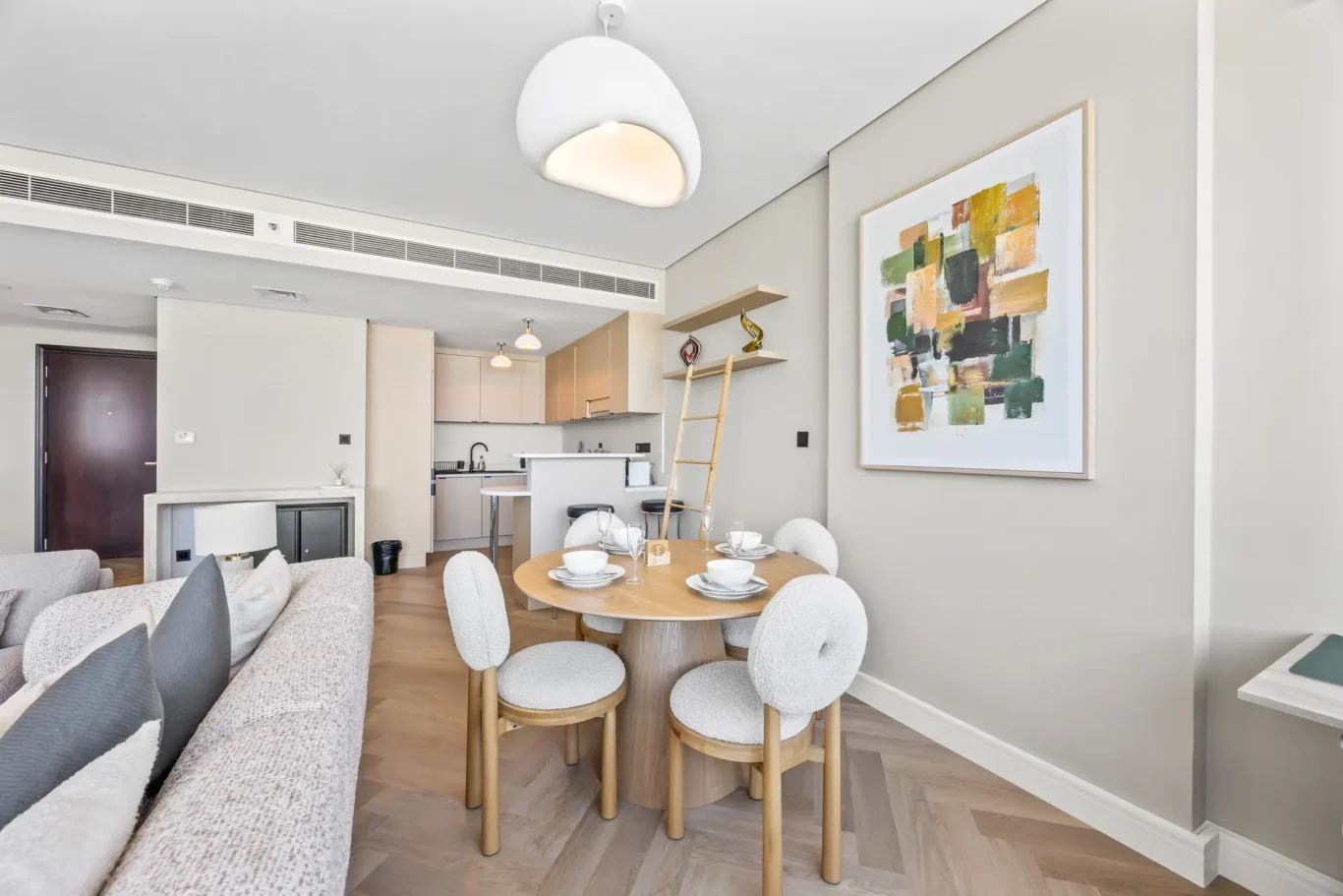 Open-plan living area with a round dining table set for six, beige chairs, a gray sofa, and a modern kitchen in the background with pendant lights showing overhead.