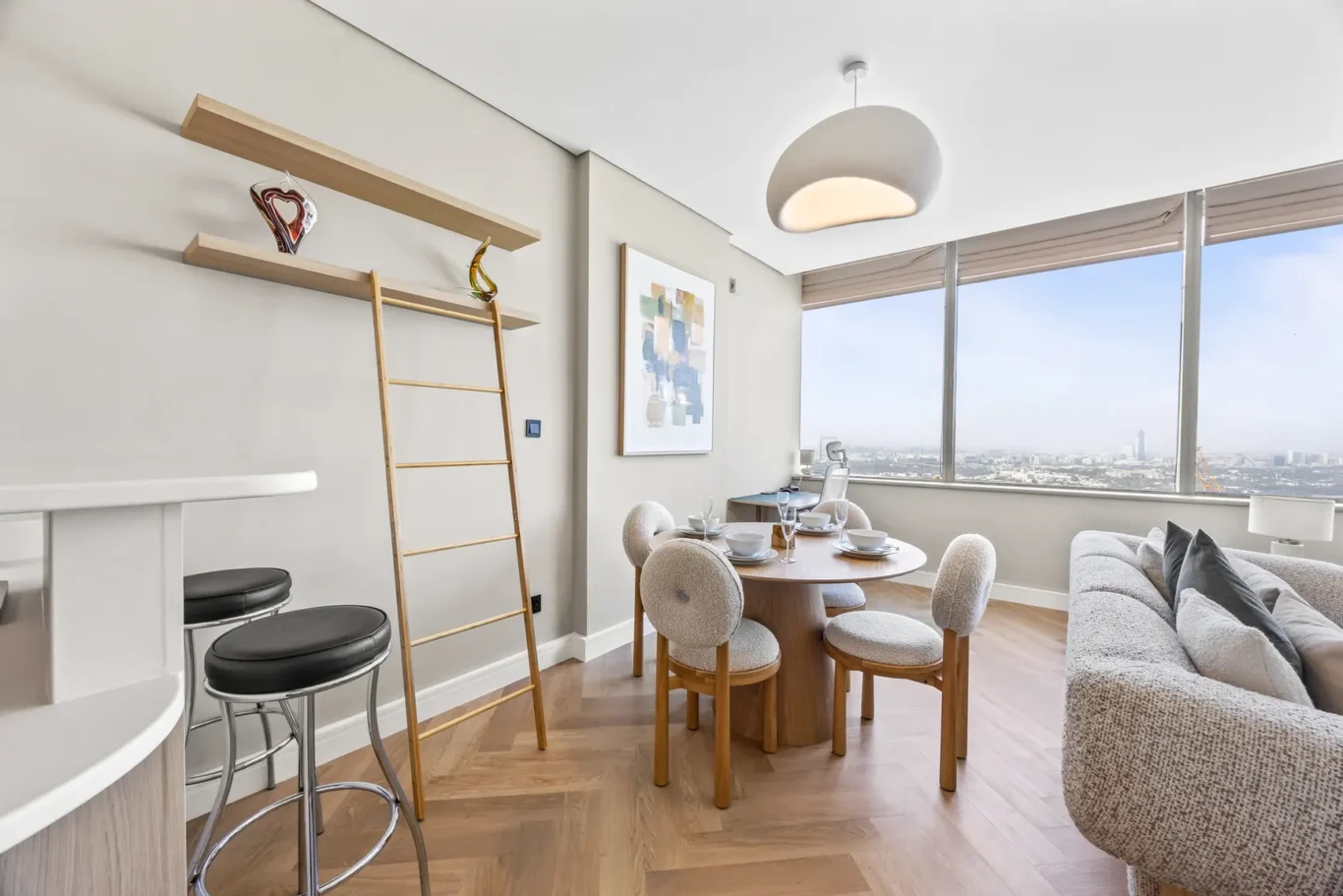 Bright living-dining area with a round wooden table set for four, plush chairs, and a large window city view.