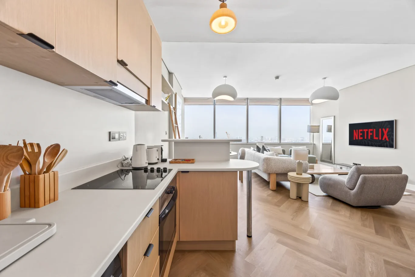 Open-plan kitchen and living area with wooden cabinets, white countertops, and a Netflix screen on the wall.