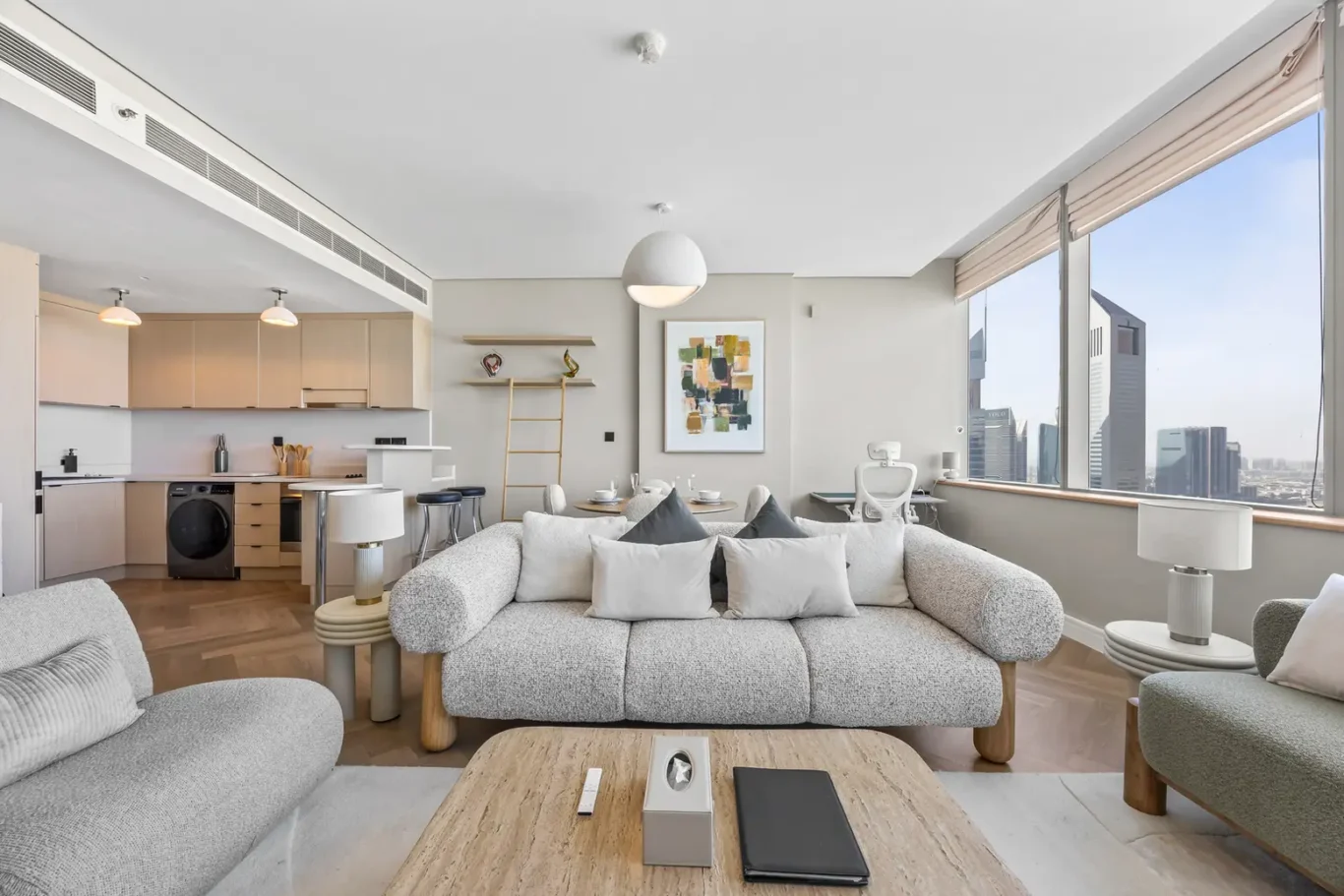Bright open-plan living room and kitchen in a high-rise apartment, featuring a gray sofa, wooden coffee table, and large city-view windows.