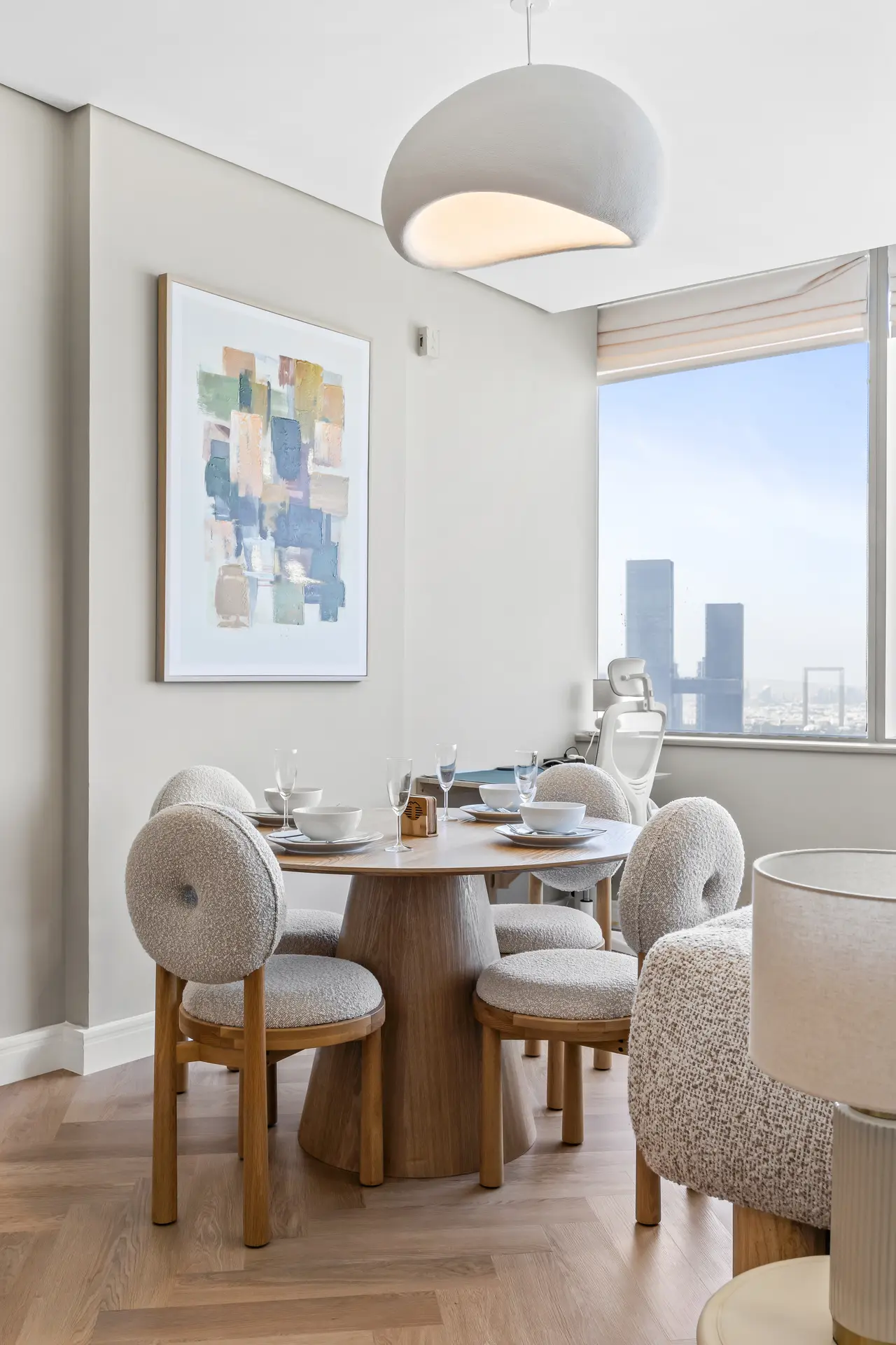 Round wooden dining table with six beige boucle chairs, abstract wall art, and a large window showing a city skyline; a modern pendant lamp hangs above.