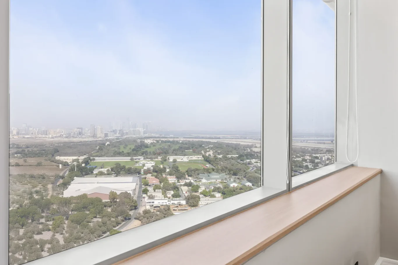 Interior window with a wooden sill overlooking a cityscape of low-rise buildings and a park outside.