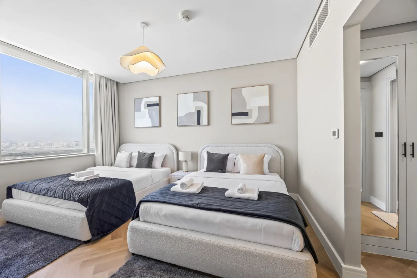 Twin beds in a bright hotel room with abstract wall art, large window overlooking the city skyline and soft gray bedding.