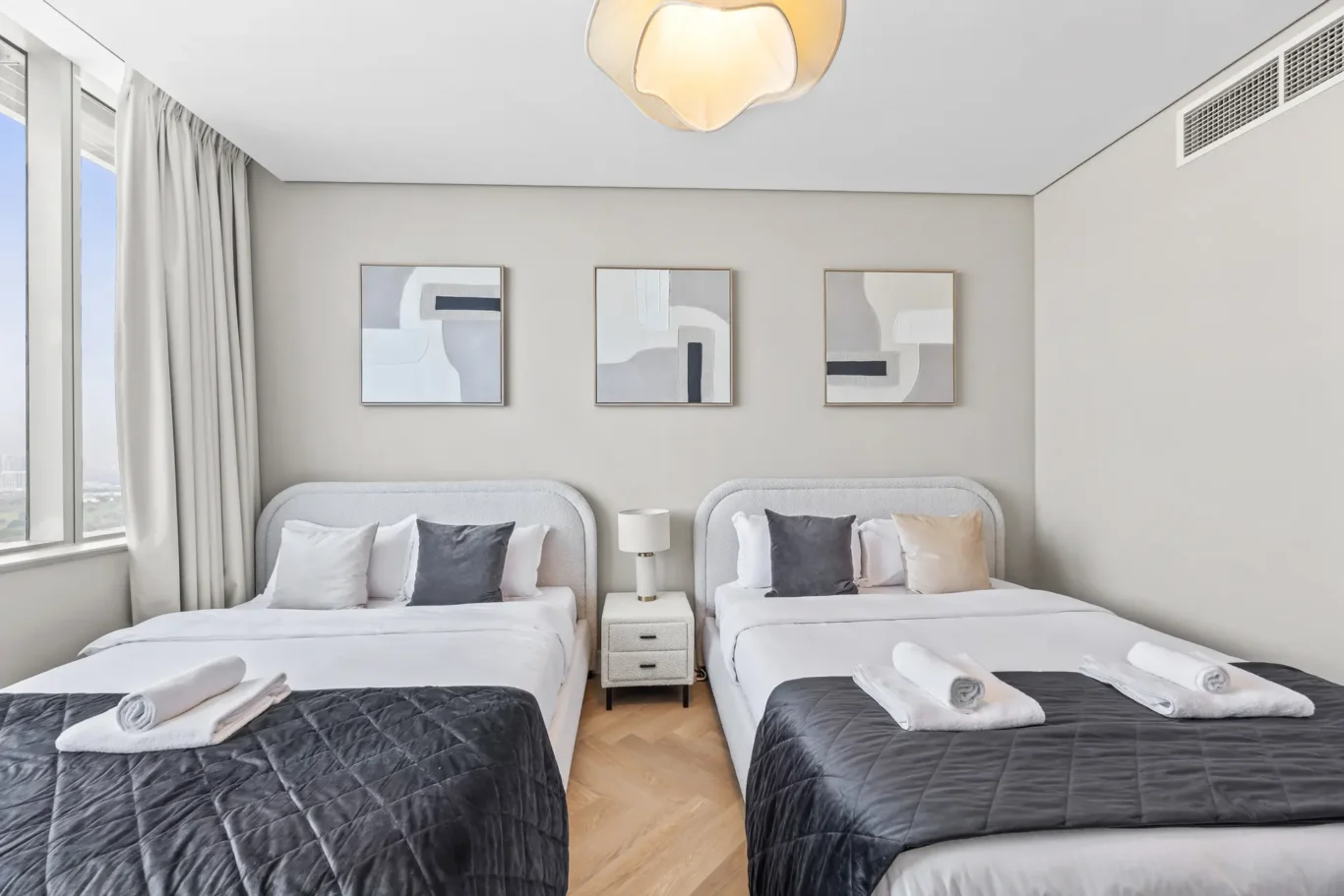 Twin hotel beds with dark quilted duvets and a shared nightstand in a bright, modern room with abstract wall art above the headboards.