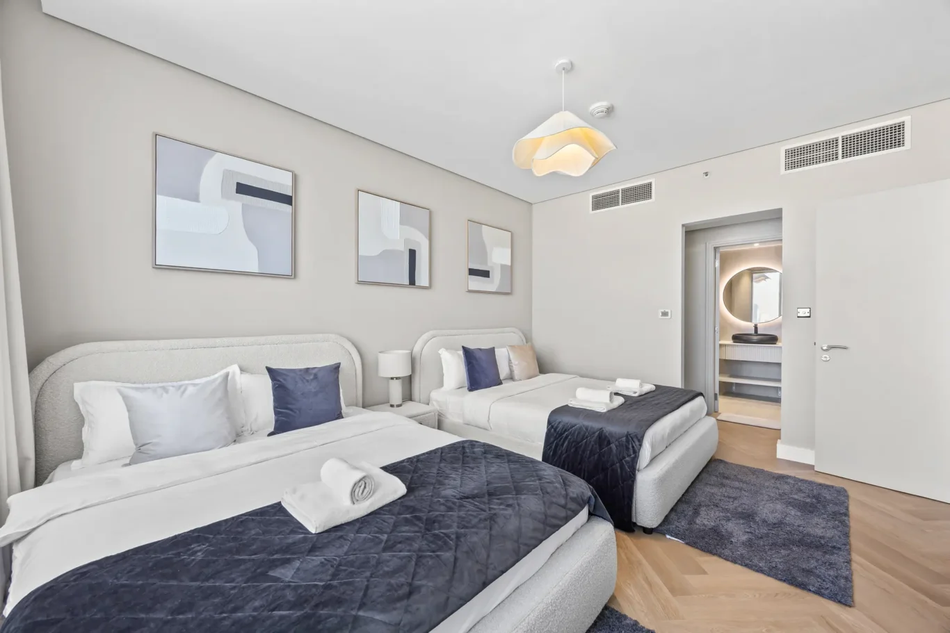Twin hotel room with two gray upholstered beds, white linens, navy throws, and folded towels, plus abstract wall art and a doorway to a bathroom with a circular backlit mirror.