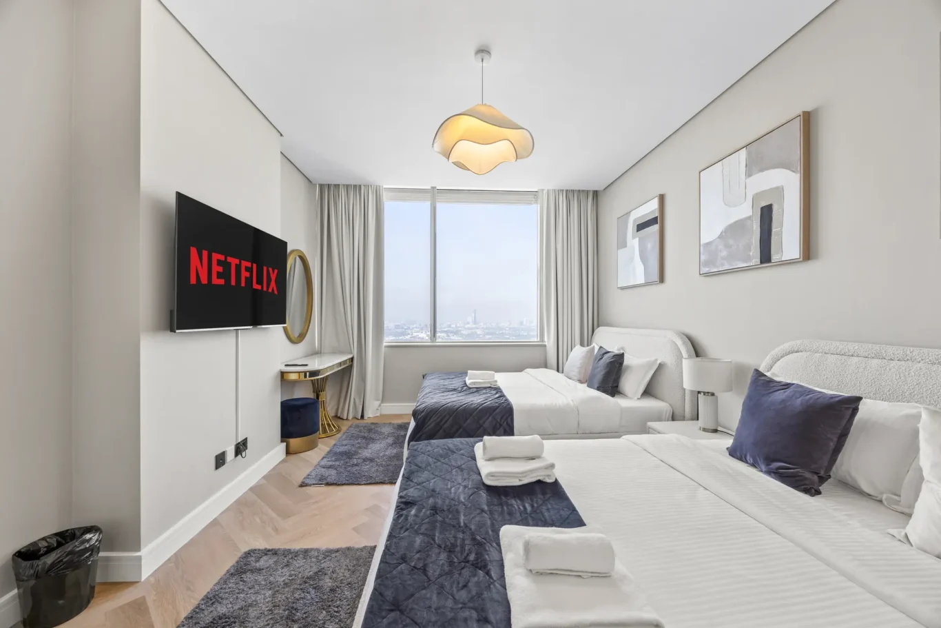 Twin hotel room with two beds, a Netflix-equipped wall TV, abstract art, and a large window with city view.