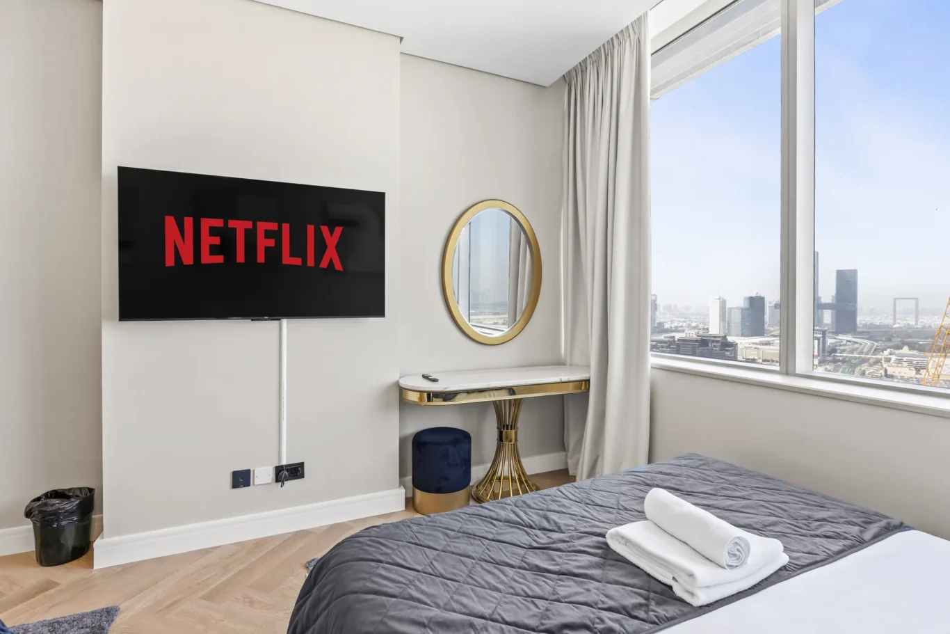 Modern bedroom with a wall-mounted Netflix screen, round gold mirror, marble desk, and a large window with a city view.