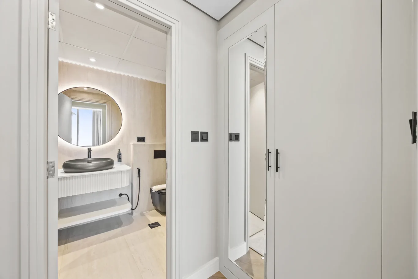 Open doorway into a modern bathroom: round black vessel sink on a white vanity, circular backlit mirror, beige tiled walls, and a wall-mounted toilet; a hallway with built-in white cabinetry and a full-length mirror on the door is visible outside the bathroom.