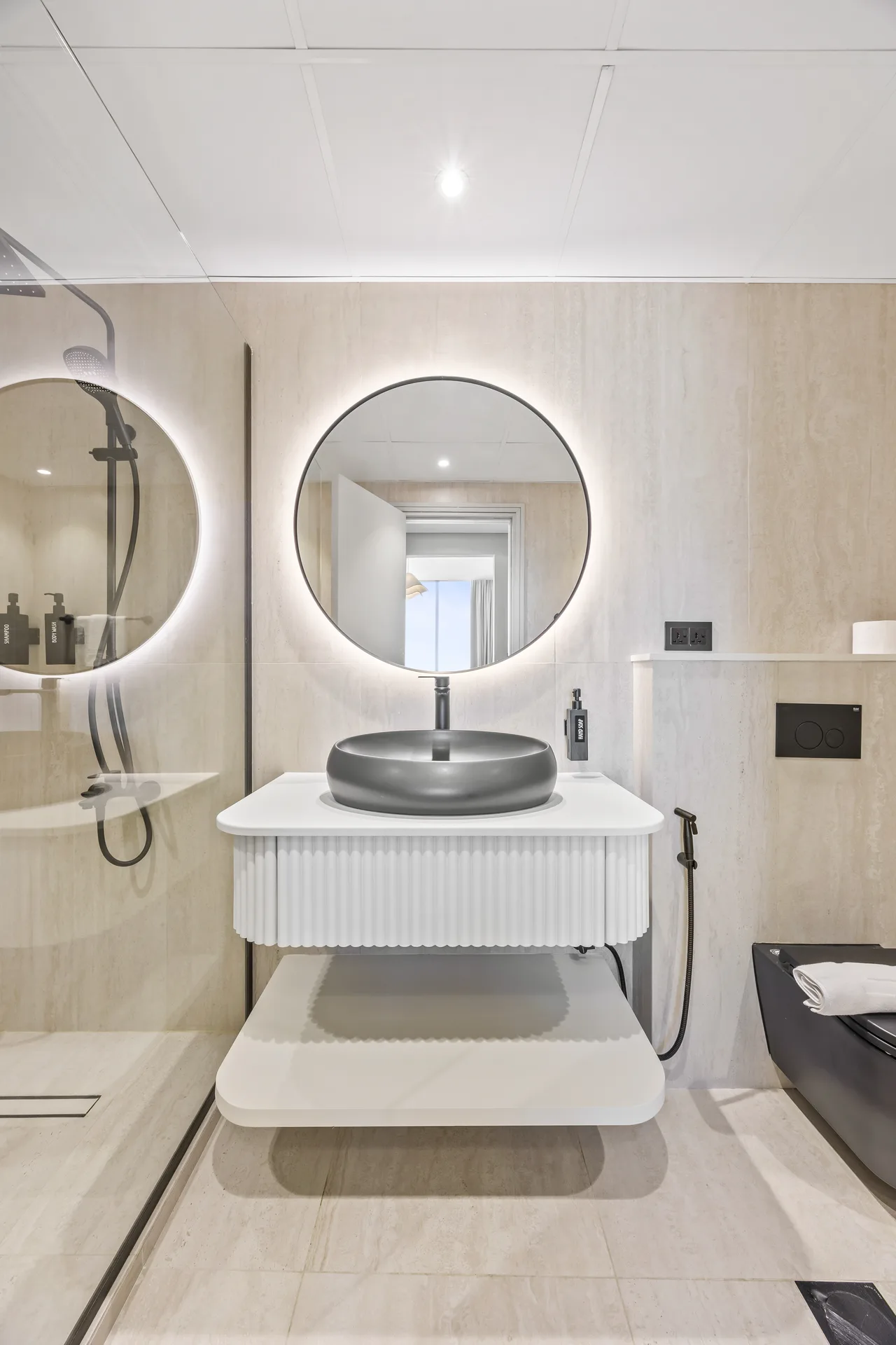 Modern bathroom vanity with a black vessel sink, round backlit mirror, and ribbed white cabinet on light tiled walls and floor, shower on the left.