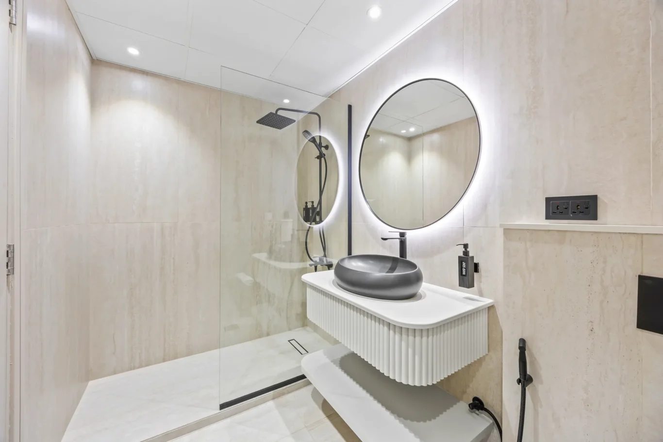 Modern bathroom with a round backlit mirror, black fixtures, and a ribbed white vanity basin behind a glass shower enclosure