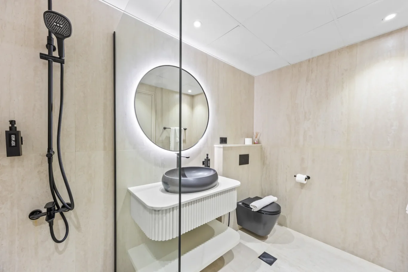 Modern bathroom with a round backlit mirror above a ribbed white vanity, a gray vessel sink, and a glass shower stall.