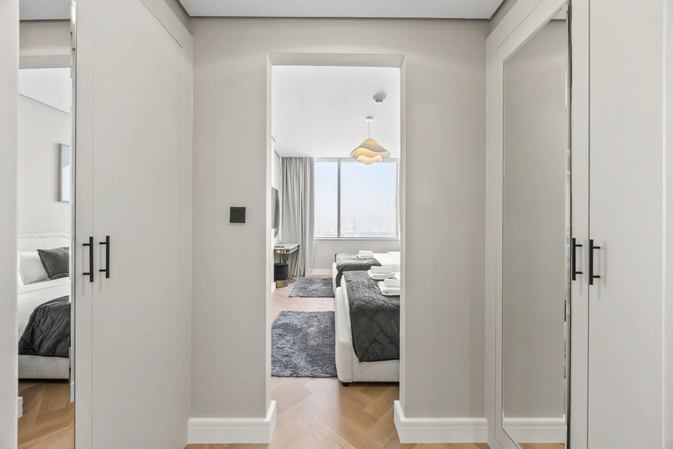 Hallway leading to a bright twin-bedroom with two beds, gray quilts, a large window, and a sculptural pendant light.