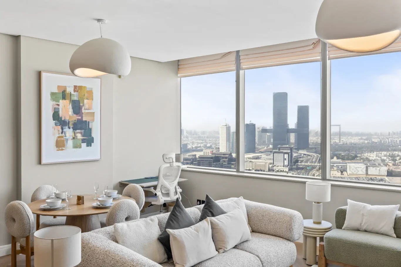 Modern living area with large windows showing a city skyline; round dining table for four and a pale sofa with cushions.