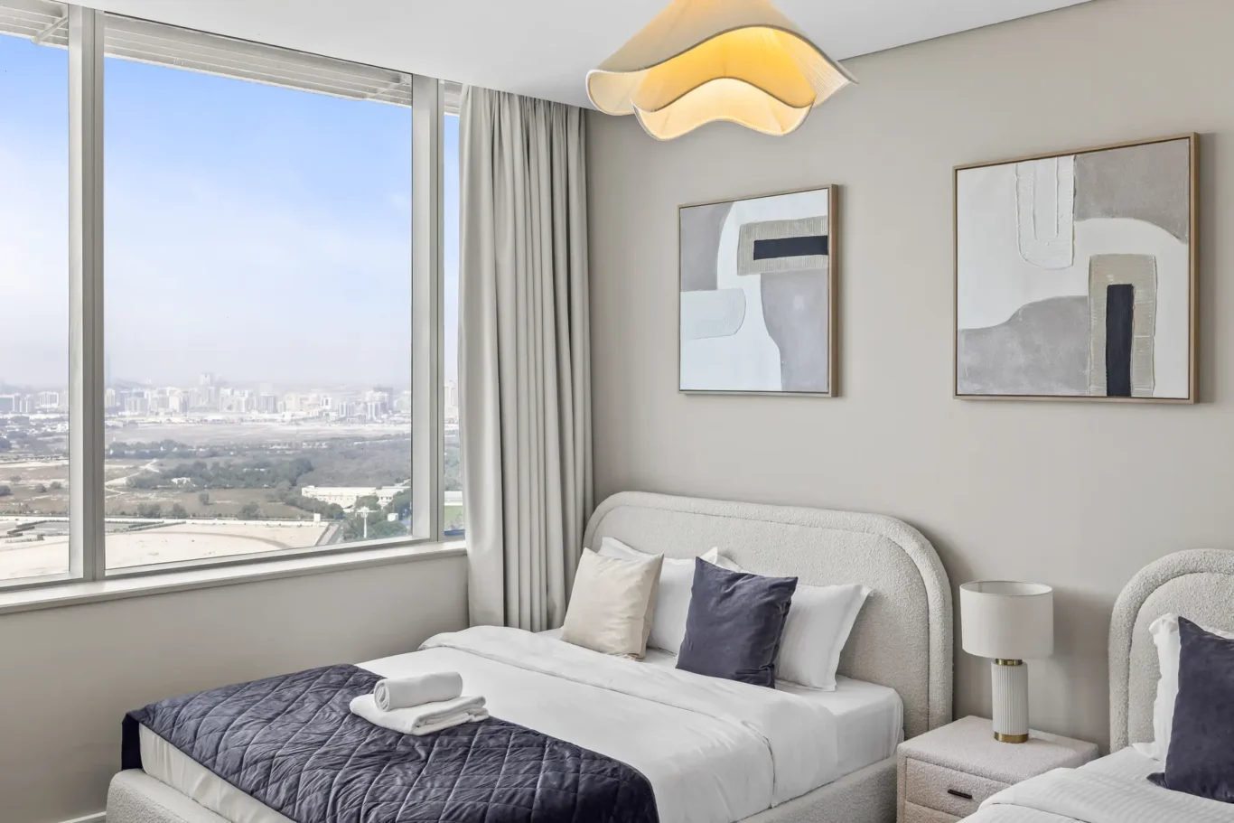 Twin bedroom with two upholstered beds, white linens, a dark blue quilt, abstract wall art, and a large window overlooking the city.