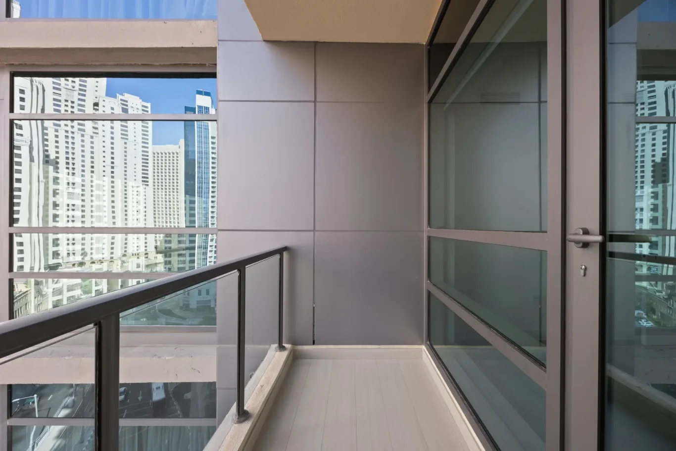 Modern balcony with glass railing on the left and large glass doors on the right; gray wall panels and light flooring, with reflected city skyscrapers and blue sky in the glass panels.