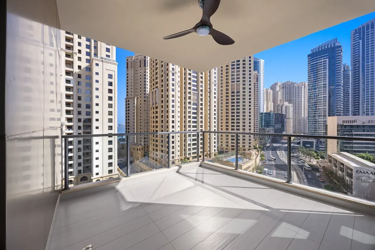 Balcony with glass railing and a ceiling fan, overlooking tall beige and glass skyscrapers under a blue sky.