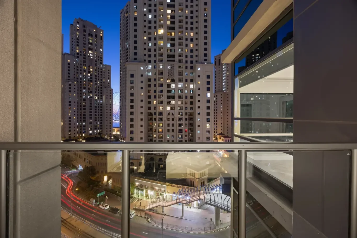 Night cityscape from a balcony: tall illuminated high-rises, blue dusk sky, and light-trail streets below.