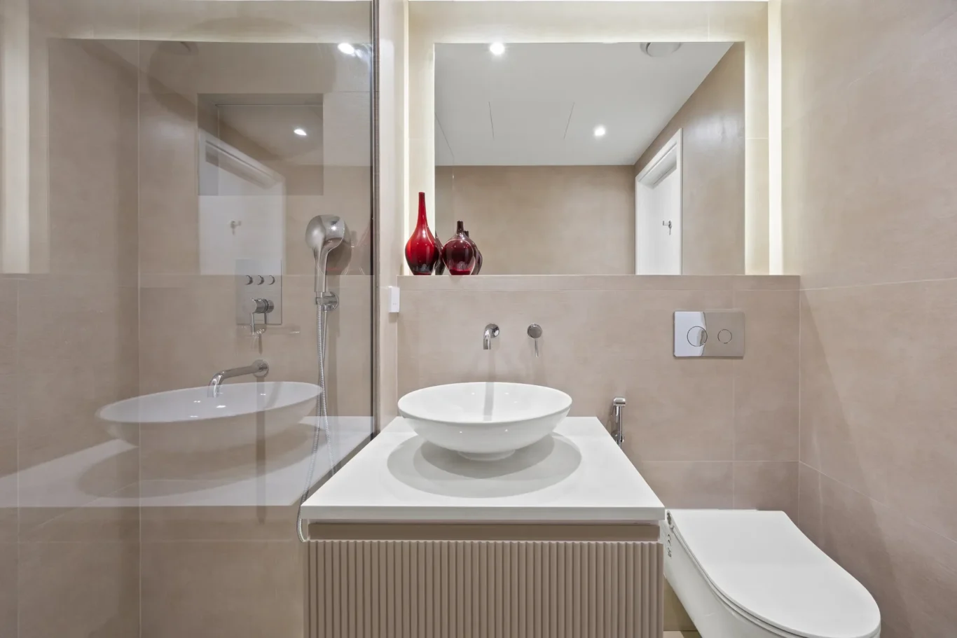 Modern beige bathroom with a glass-walled shower, white vessel sink, and wall-mounted controls; two red glass vases on the shelf above the sink.