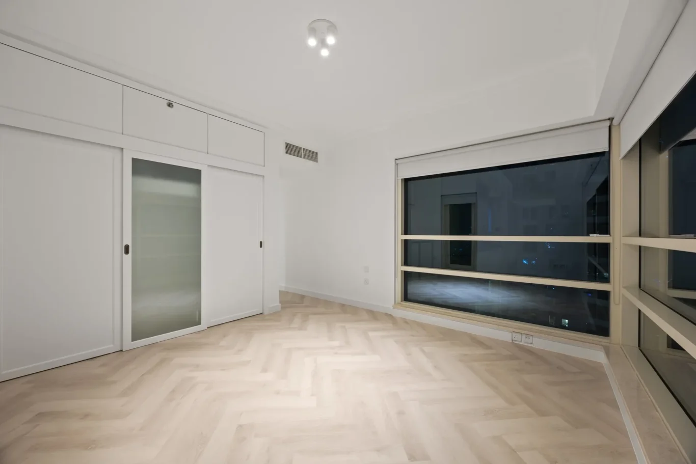 Empty, modern room with white built-in cabinets and frosted glass door along left wall, a large window on the right, and light wood herringbone flooring at night.