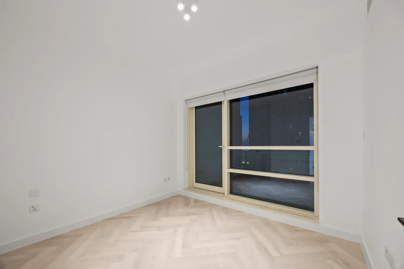 Empty modern room with a large window, white walls, and a light herringbone wood floor. Evening outside visible through the window.