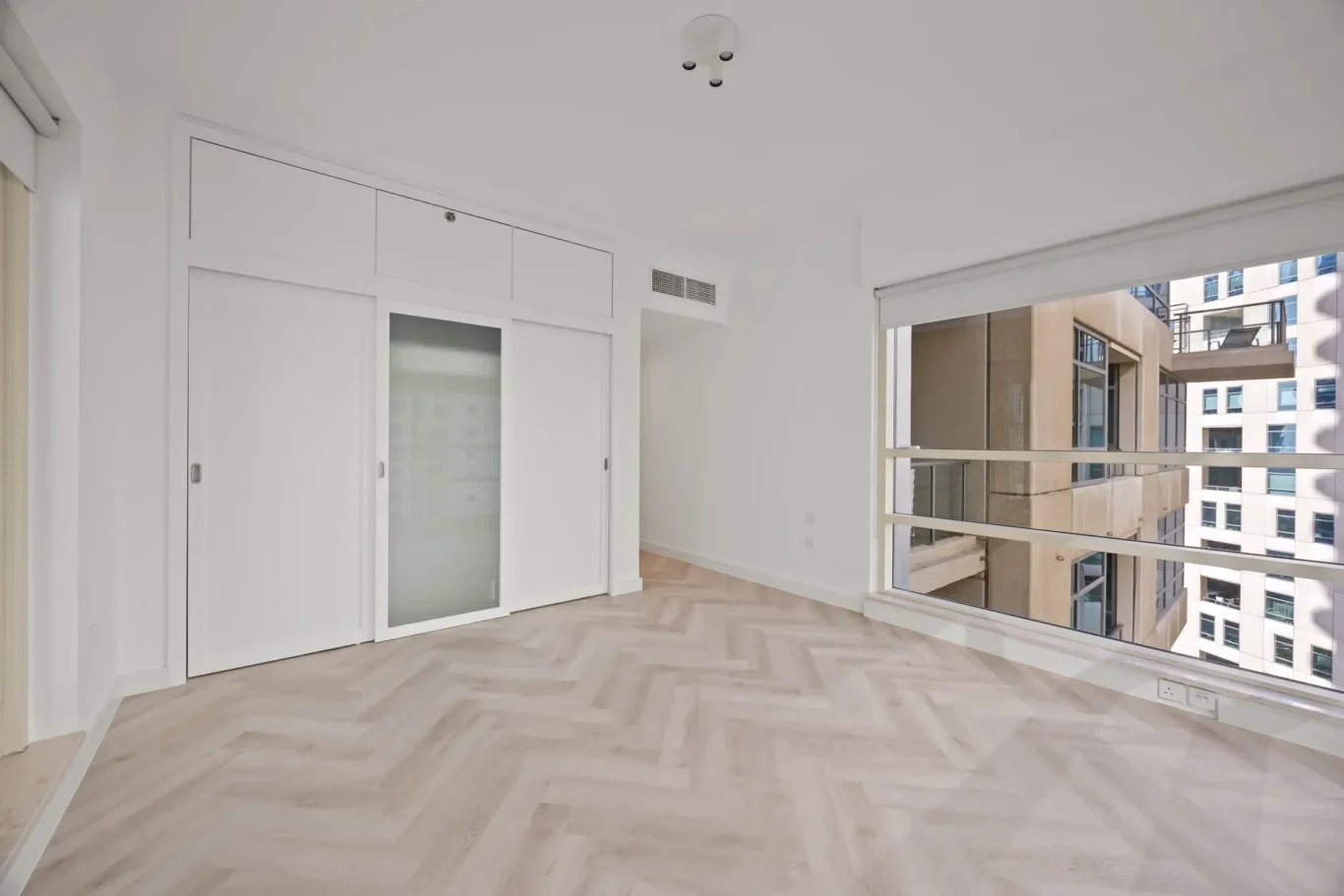 Bright minimalist living room with white walls, built-in storage, and a large window opening to a balcony overlooking city buildings.