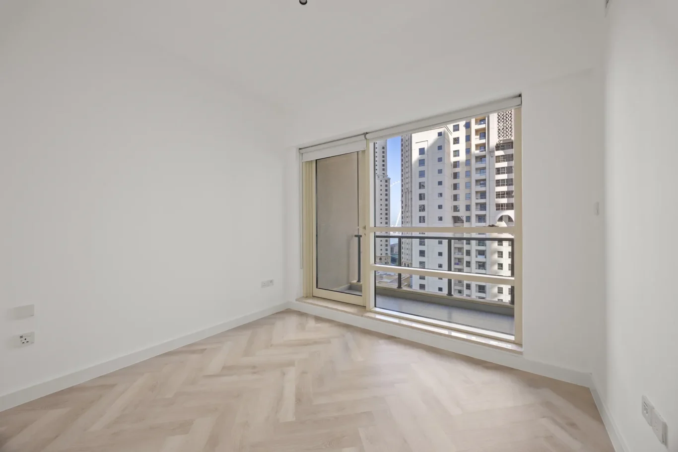 Bright empty room with white walls, light wood floor, and a large sliding glass door opening to a balcony with city buildings outside.