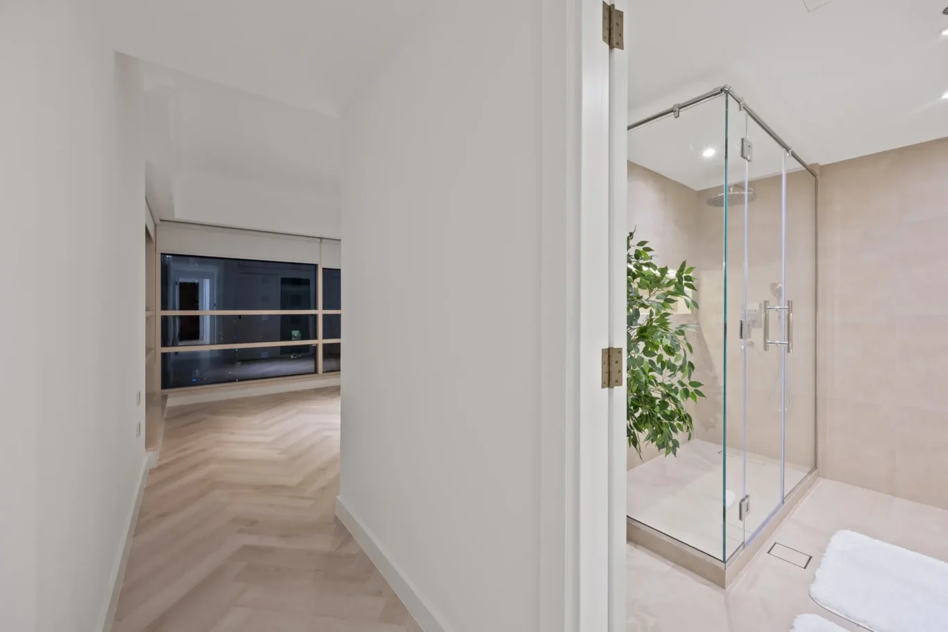 Bright hallway with light wood floors leading to a living area; glass-enclosed shower visible on the right side.