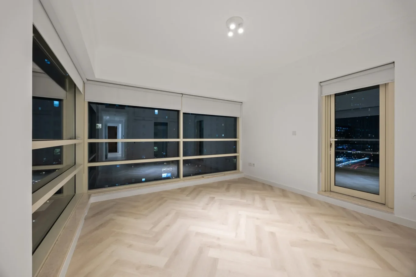 Bright empty living room with large floor-to-ceiling windows and a balcony door, city lights visible at night on the horizon.
