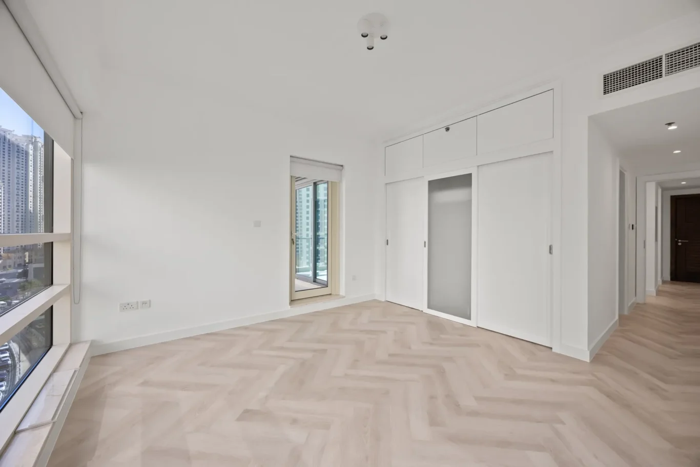 Bright, empty living area with large windows on the left, light wood herringbone flooring, and white walls. A built-in closet with sliding doors is on the far wall.