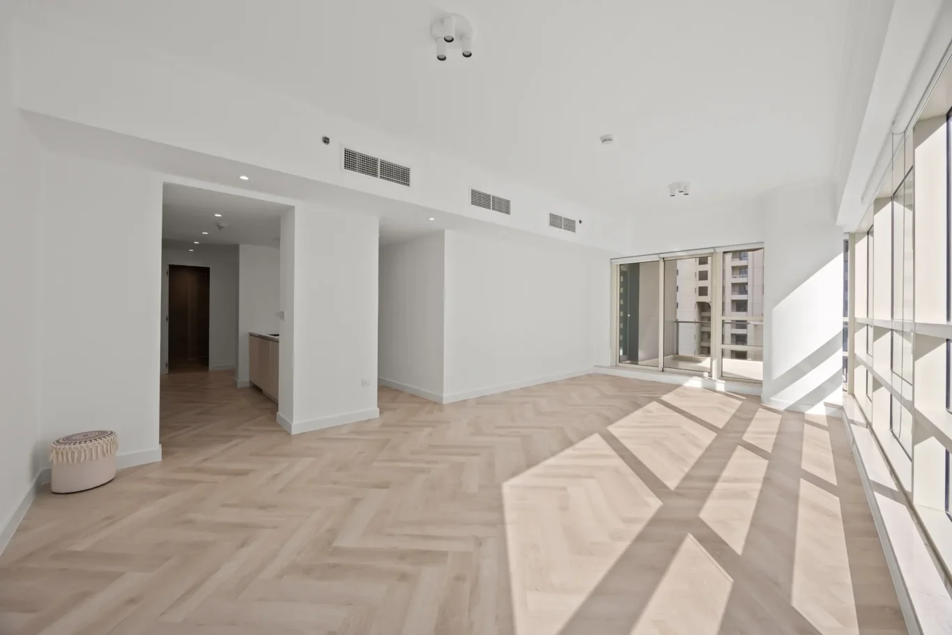 Bright open-plan living space with white walls, large floor-to-ceiling windows, and light wood herringbone flooring on a sunny day.