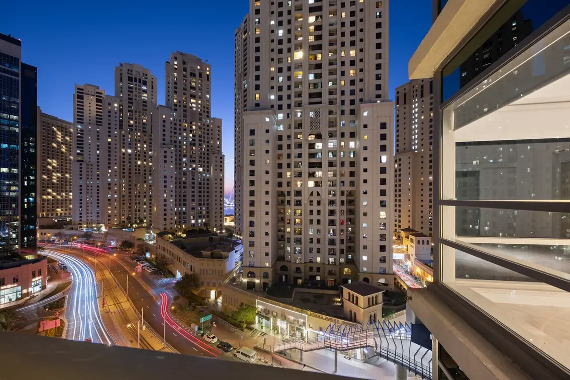 Night cityscape of tall beige apartment towers with lit windows, curved light-trail streets, and a glass-fronted building on the right.