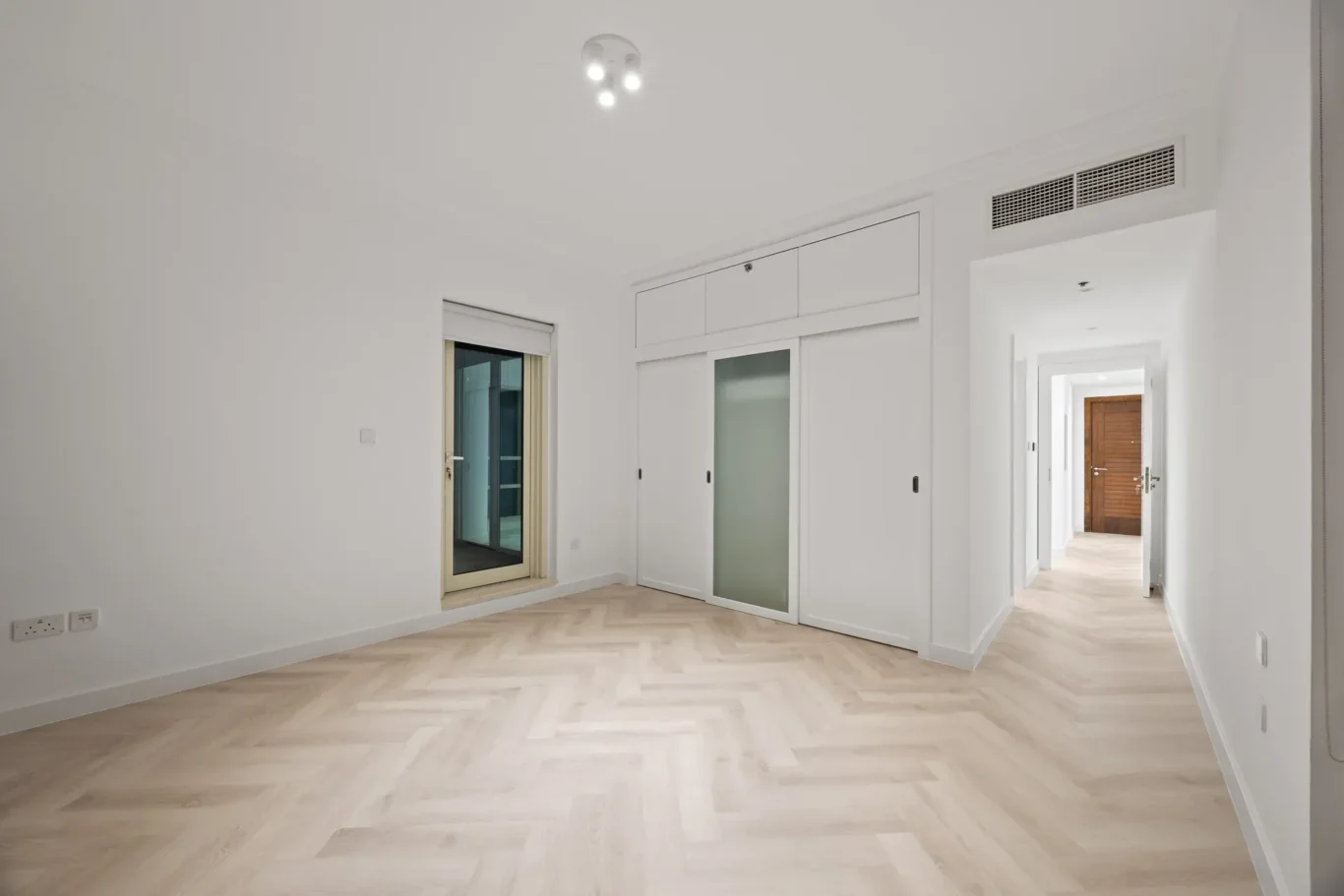 Empty white room with a pale wood herringbone floor, built-in white wardrobes, and a hallway leading to a wooden door.