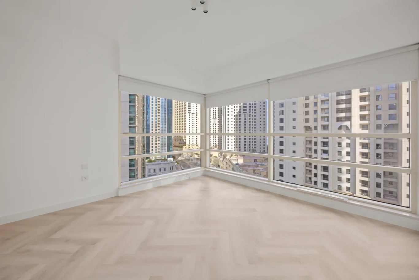 Bright empty corner room with large wraparound windows showing high-rise cityscape outside, light wood floor, white walls, and roller blinds partially up.
