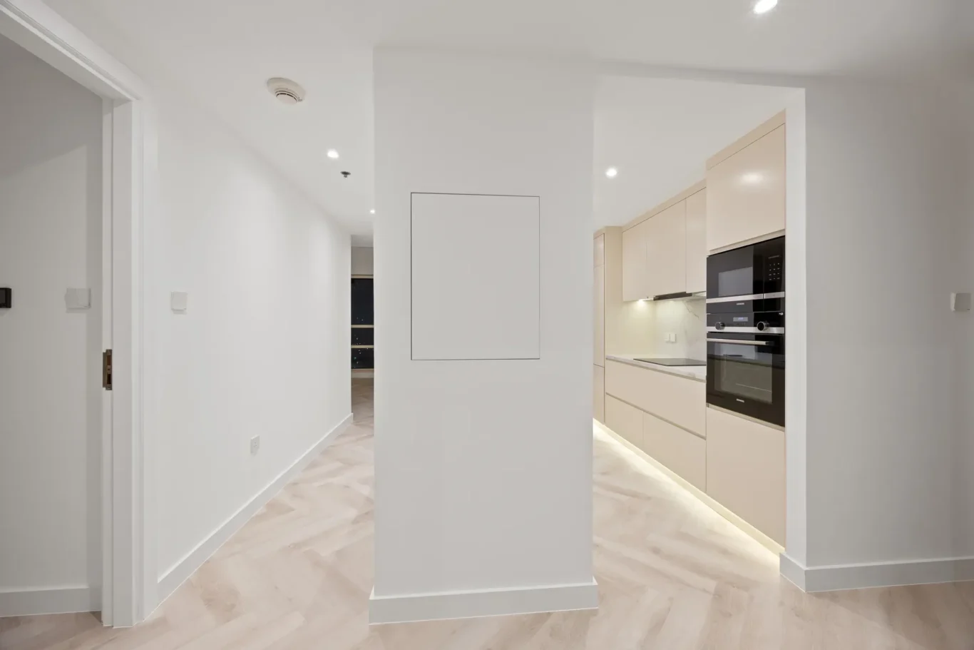 Open-plan hallway leading to a modern beige kitchen with built-in oven and under-cabinet lighting.