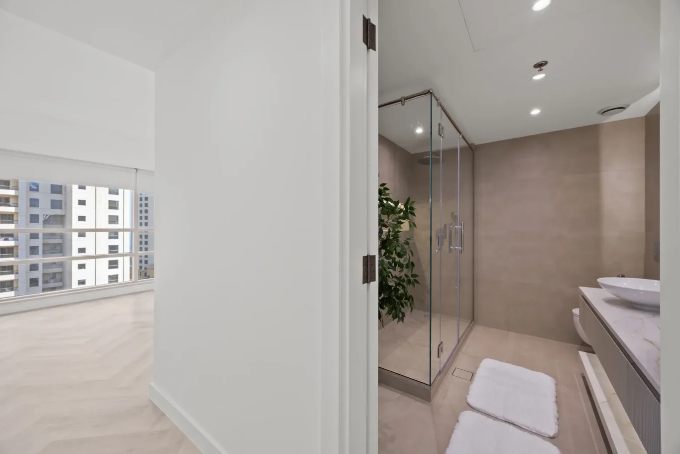 Modern bathroom with a glass-enclosed shower, beige tiled walls, and a white vessel sink with a long vanity; two white bath mats on the floor and a green plant in the corner.