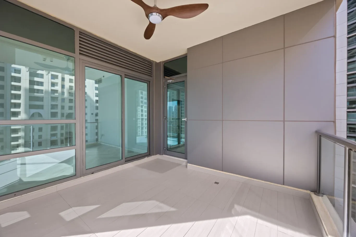 Empty balcony with large glass doors, a wooden ceiling fan, and gray panel walls overlooking neighboring high-rise buildings.