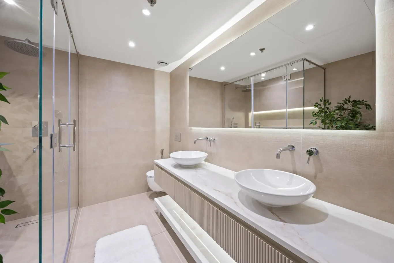 Modern beige-tiled bathroom with a double sink vanity, round vessel sinks, and a large illuminated mirror above a marble countertop.