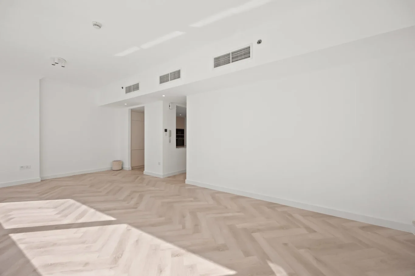 Bright empty living room with white walls, light herringbone wood floor, and ceiling vents casting geometric shadows from sunlight.