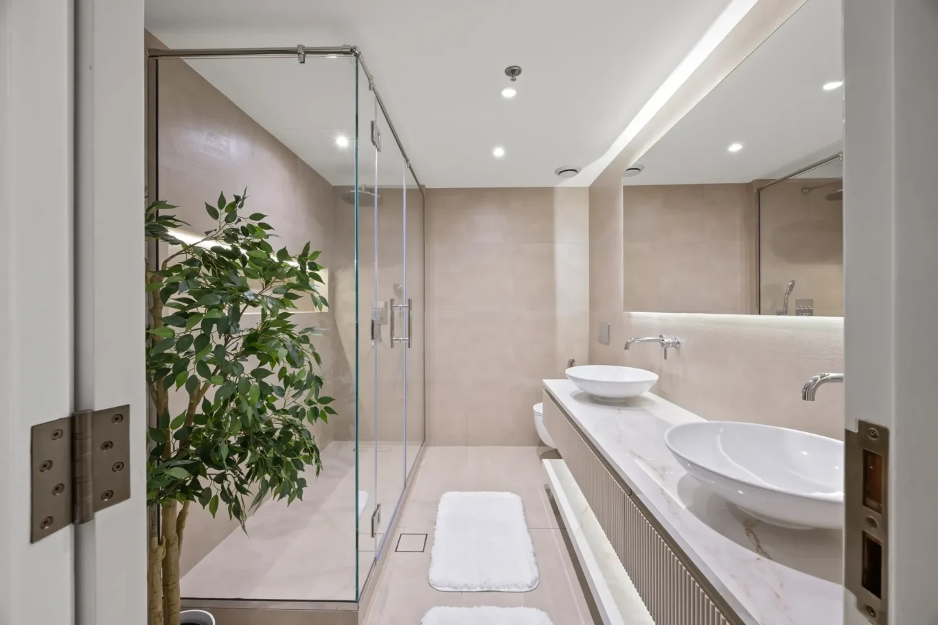 Modern bathroom with glass shower enclosure, beige tiles, white countertops, two vessel sinks, and a tall indoor plant.