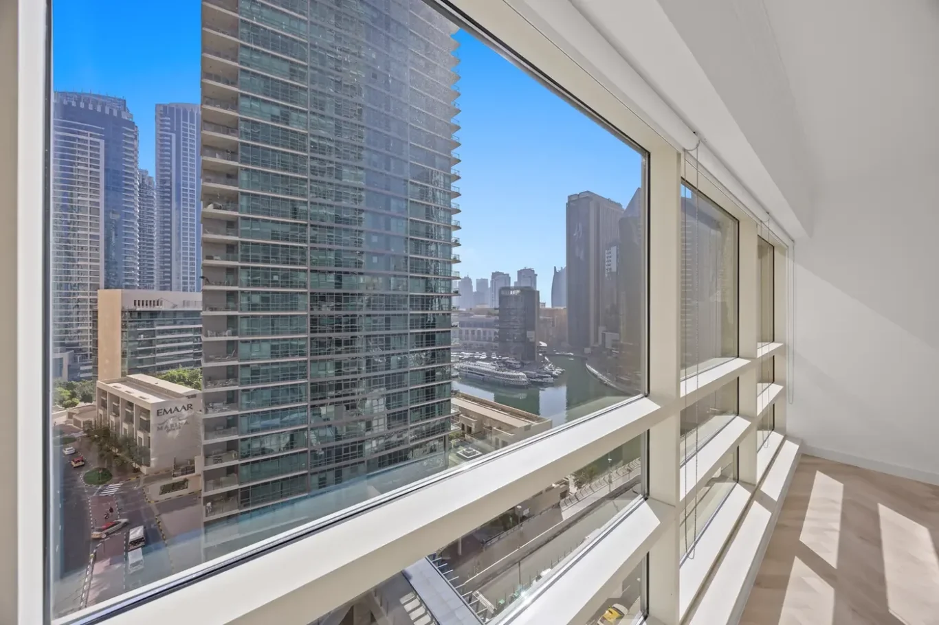 Bright, modern apartment interior with floor-to-ceiling windows overlooking a marina and tall glass skyscrapers against a clear blue sky.