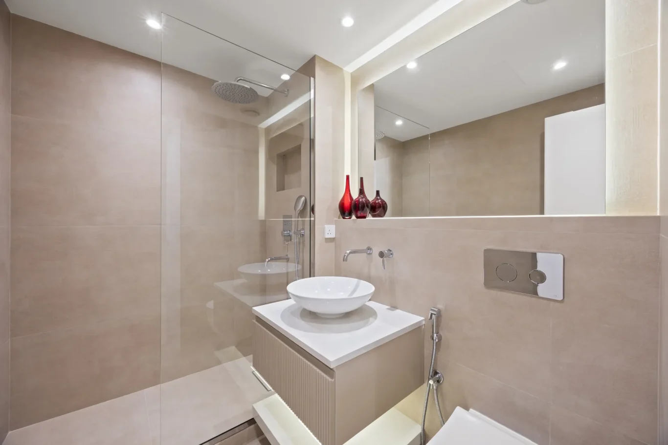 Modern bathroom with a glass shower enclosure, beige tiled walls, a white vessel sink on a beige vanity, and three red vases on the counter.
