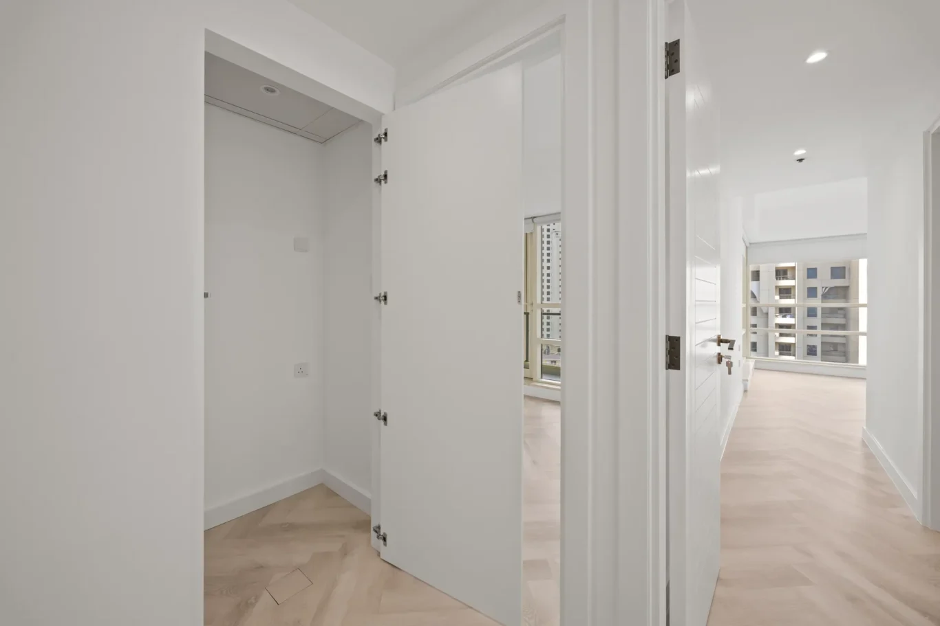 Bright, white hallway with open doors to a sunlit living area and balcony, light wood herringbone flooring.