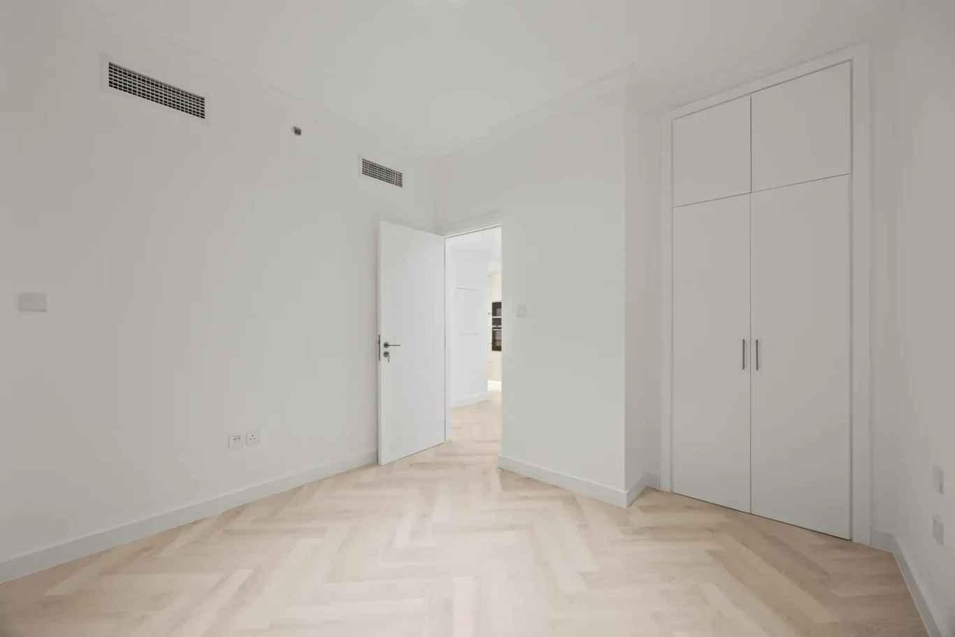 Bright white hallway with light wood chevron flooring, open door to a connected room, and a tall white closet on the right.