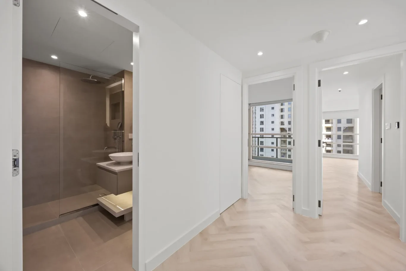 Bright hallway in a modern apartment; open bathroom is visible on the left with brown tile walls, and city buildings are seen through the windows ahead.