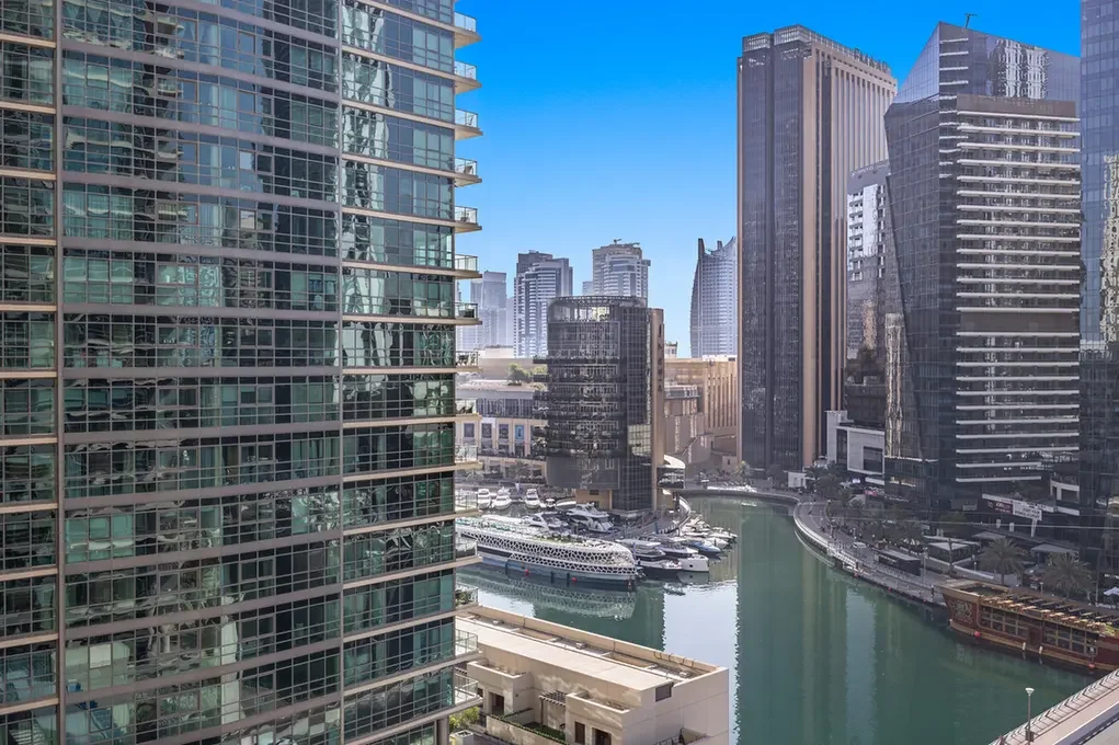 Tall glass-clad skyscrapers line a marina filled with luxury yachts under a clear blue sky, forming a modern urban waterfront.