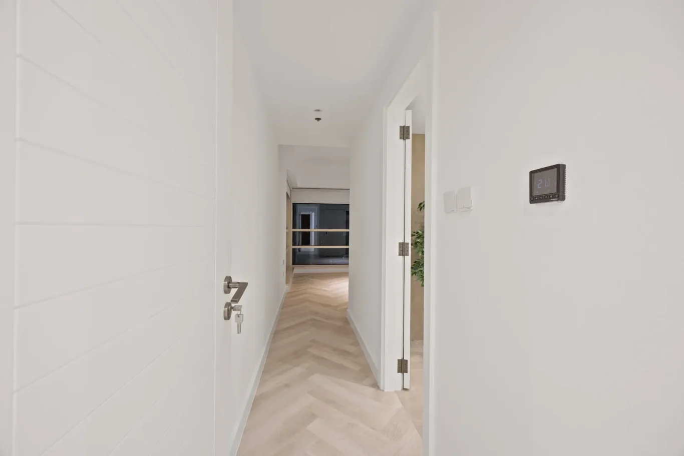 Bright white hallway with light wood chevron flooring, several open doors, and a wall thermostat on the right wall.