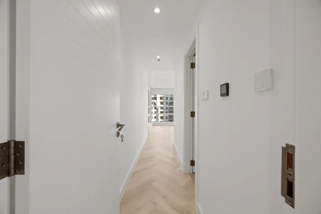 Bright white hallway with doors along both sides, leading to a sunlit room at the end and a light wood chevron floor.