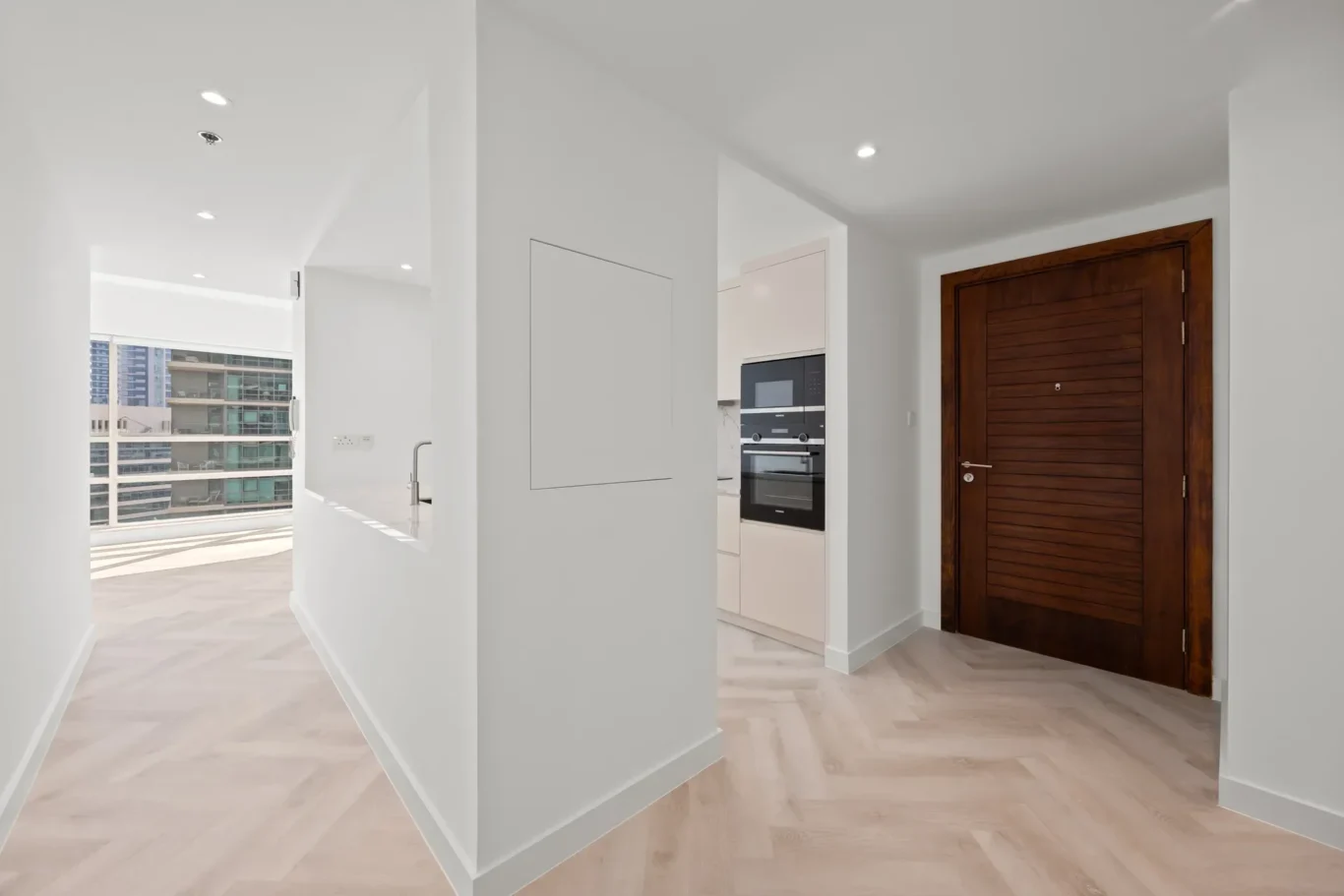 Modern white hallway leading to a kitchen with built-in oven; dark wooden entry door on the right era.