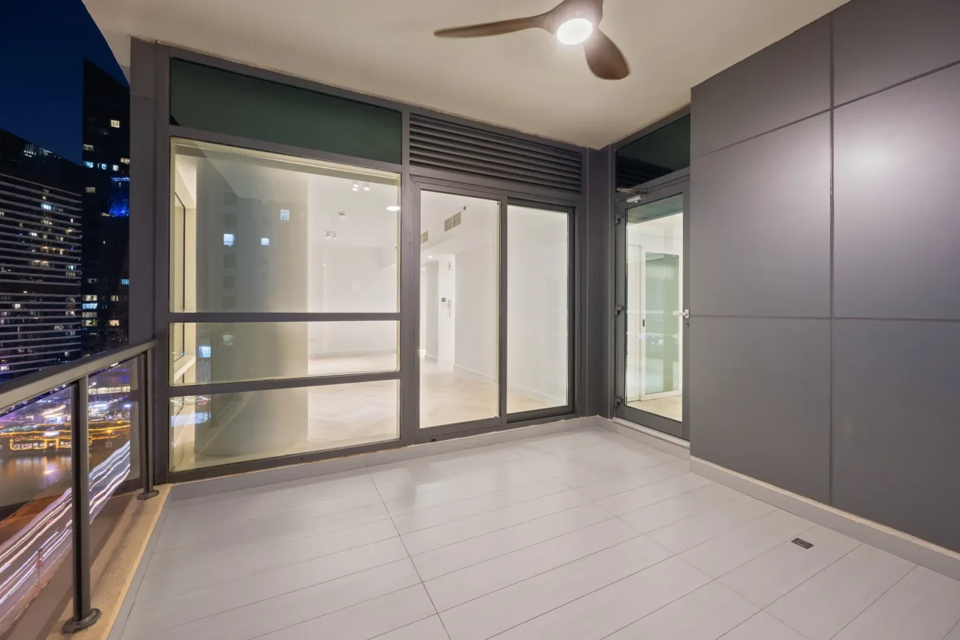 Balcony with glass doors overlooking city lights at night; white interior room beyond, gray walls, and a ceiling fan overhead.