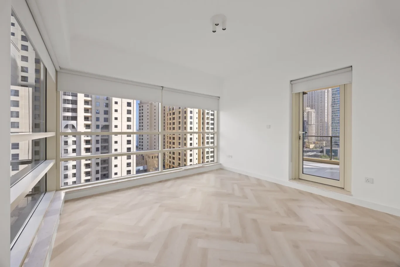 Bright empty living area with large windows and a balcony door showing city high-rises outside.